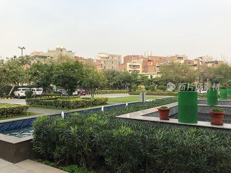 印度新德里公园的现代现代花园设计形象，有喷泉线，正式的蓝色马赛克瓷砖反射池/池塘，修剪树篱和传统的印度房屋